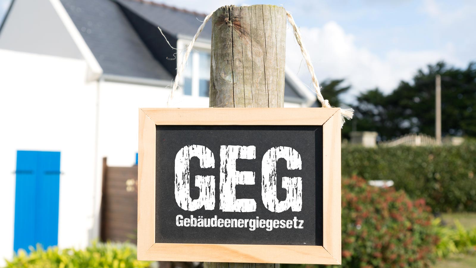 Holzpfahl mit einen Schild und der Aufschrift GEG Gebäudeenergiegesetz vor einem Einfamilienhaus im Hintergrund