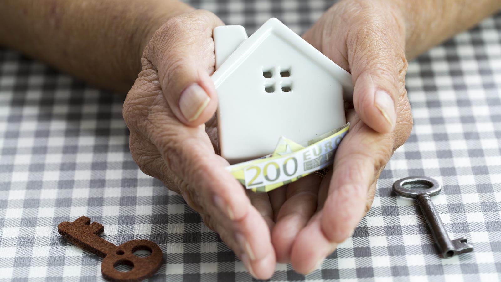 Zwei Hände einer älteren Person halten ein Modellhaus und Geldschein über einen Tisch mit zwei Schlüsseln