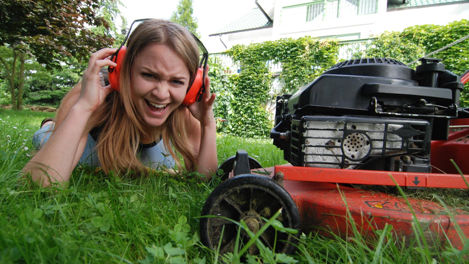Von Lärm genervte Frau mit Kopfhörern liegend neben einem Rasenmäher