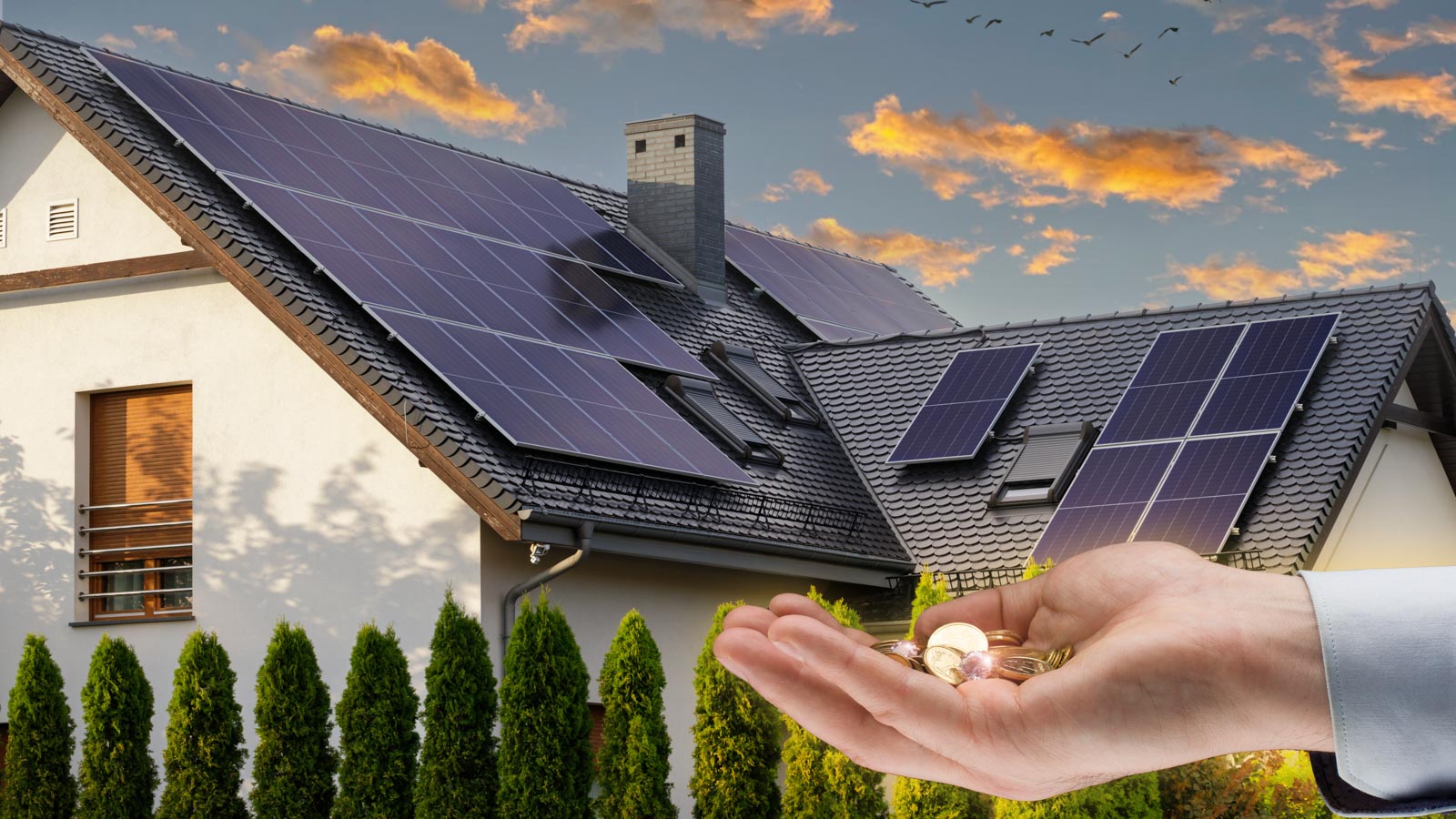 Hand mit Geldmüzen im Vordergrund vor einem Haus mit Photovoltaik-Anlage