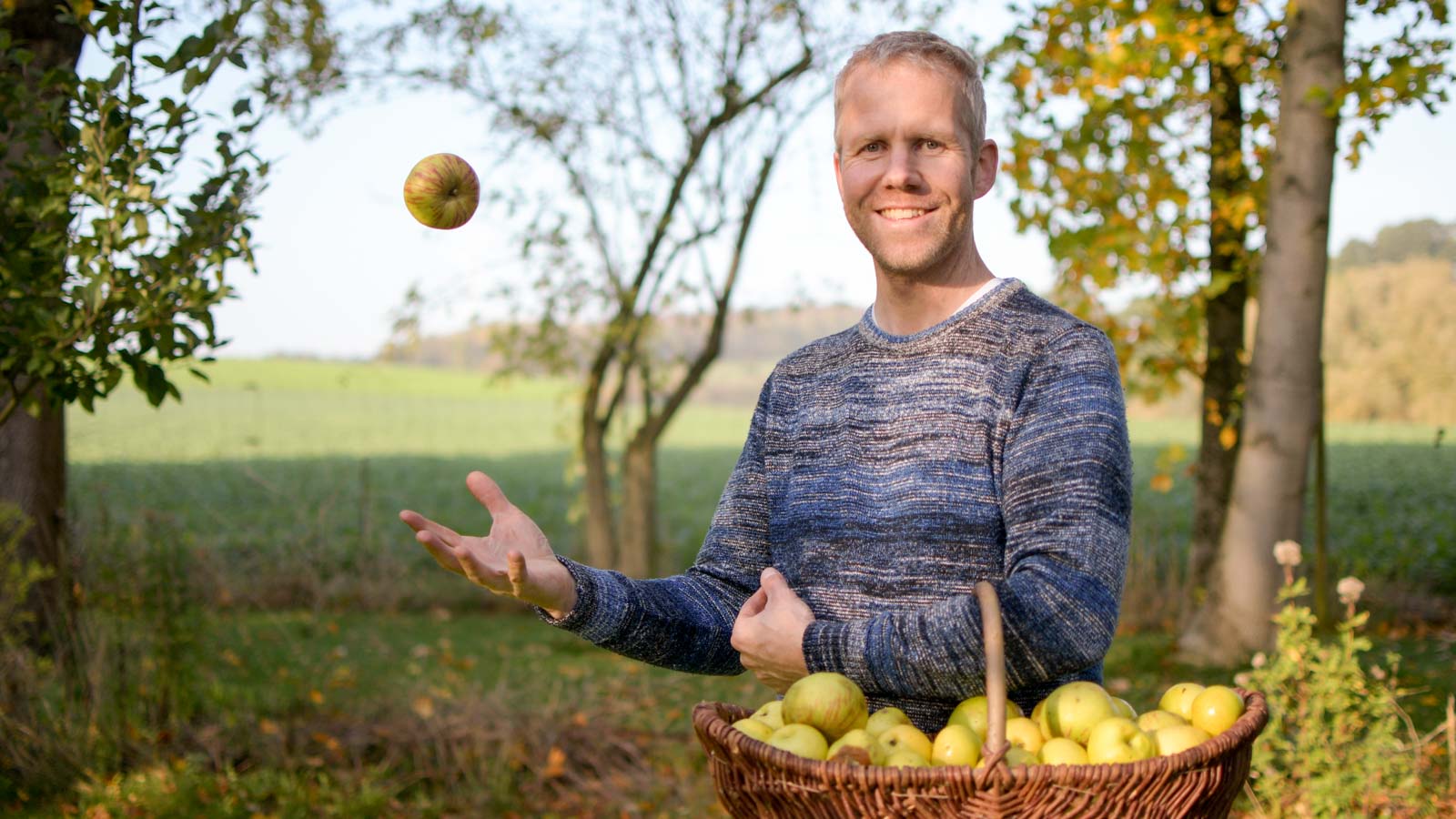 Gartenberater Philippe Dahlmann steht im Garten, hat einen Korb voller Äpfel im Arm und wirft einen Apfel in die Luft.