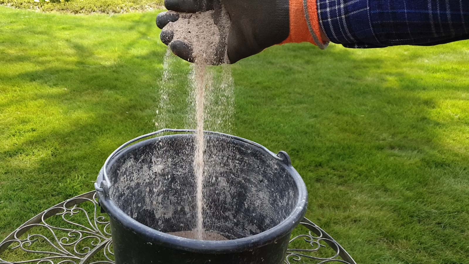 Gewaschener Rheinsand 0/2 rieselt durch eine Hand in einen Eimer. Im Hintergund ein Rasen der noch vertikutiert wurde.