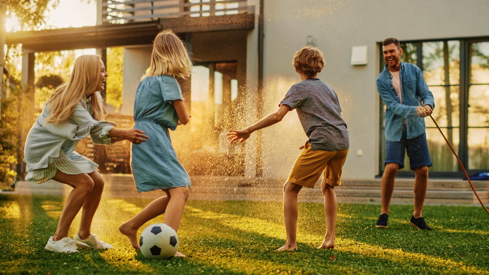 Eltern toben mit ihren Teenager-Kindern an einem sommerlichen Spätnachmittag im Garten mit Fußball und Gartenschlauch. 