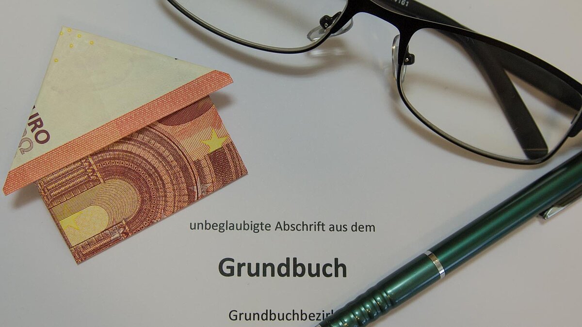Zum Haus gefalteter Euroschein, Brille, Kugelschreiber und Blatt mit dem Aufdruck Grundbuch
