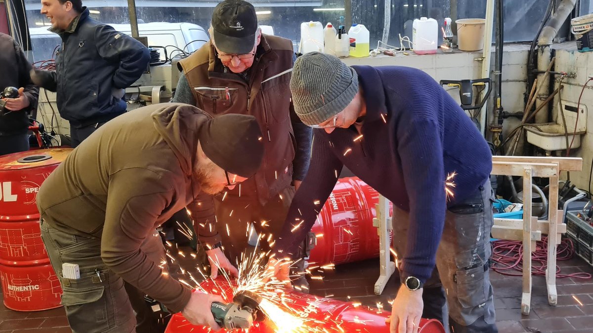 In einer Werkstatt arbeiten drei Männer an einem Stahlfass mit einem Flex-Winkelschleifer