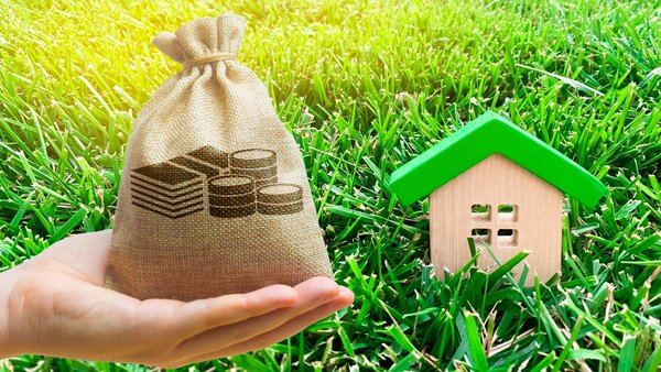 Auf dem Rasen ist ein Holzhaus, im Vordergrund ist eine Hand die ein Sack voll Geld in der Hand hält