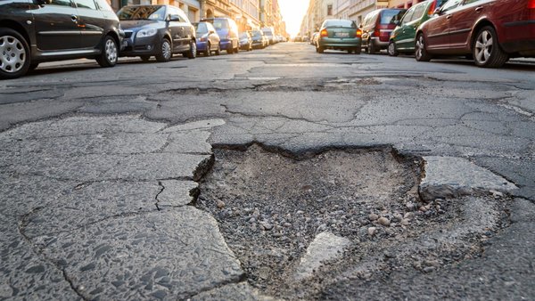 Das Bild zeigt eine von historischen Häusern und parkenden Autos gesäumte Straße beim Sonnenaufgang. In der Mitte befindet sich ein großes Schlagloch. Die Fahrbahn ist insgesamt rissig und beschädigt.