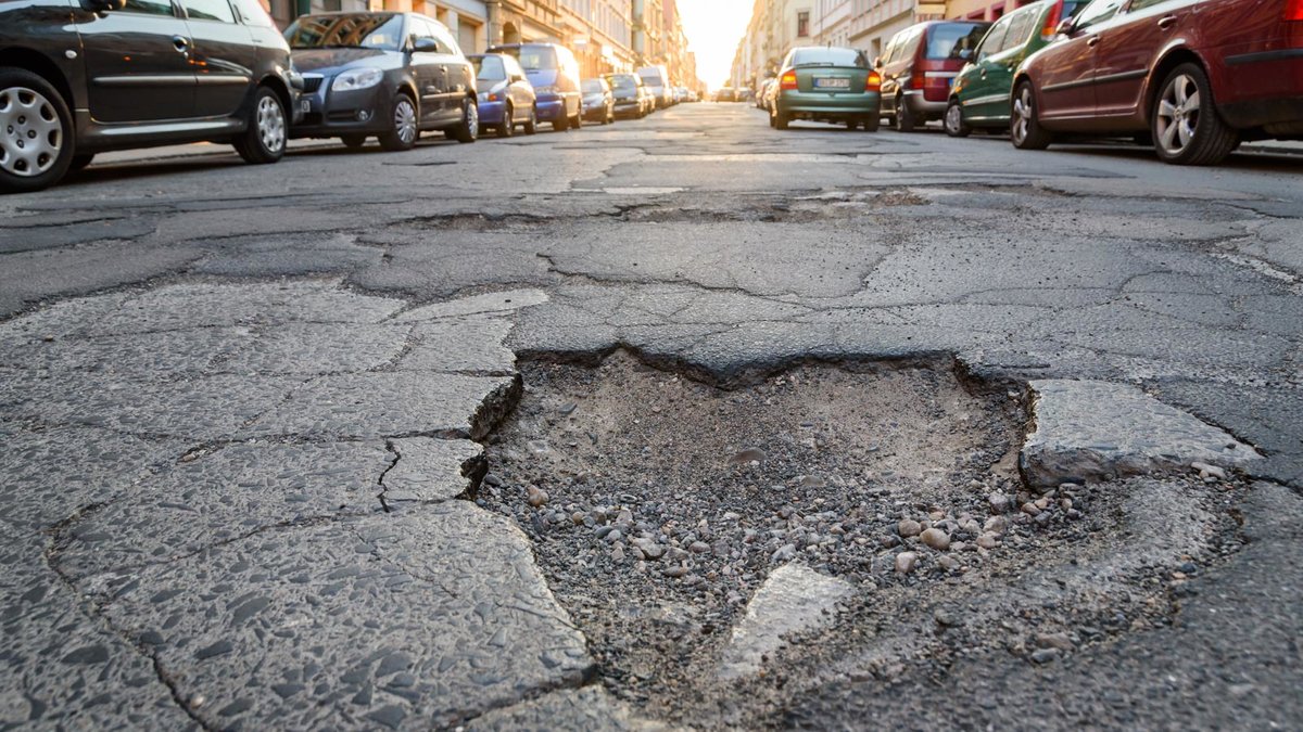 Das Bild zeigt eine von historischen Häusern und parkenden Autos gesäumte Straße beim Sonnenaufgang. In der Mitte befindet sich ein großes Schlagloch. Die Fahrbahn ist insgesamt rissig und beschädigt.