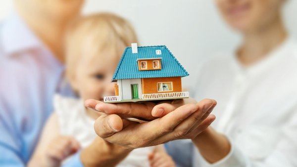 Junge Eltern mit Kleinkind halten ein Miniaturhaus auf den Handflächen. 