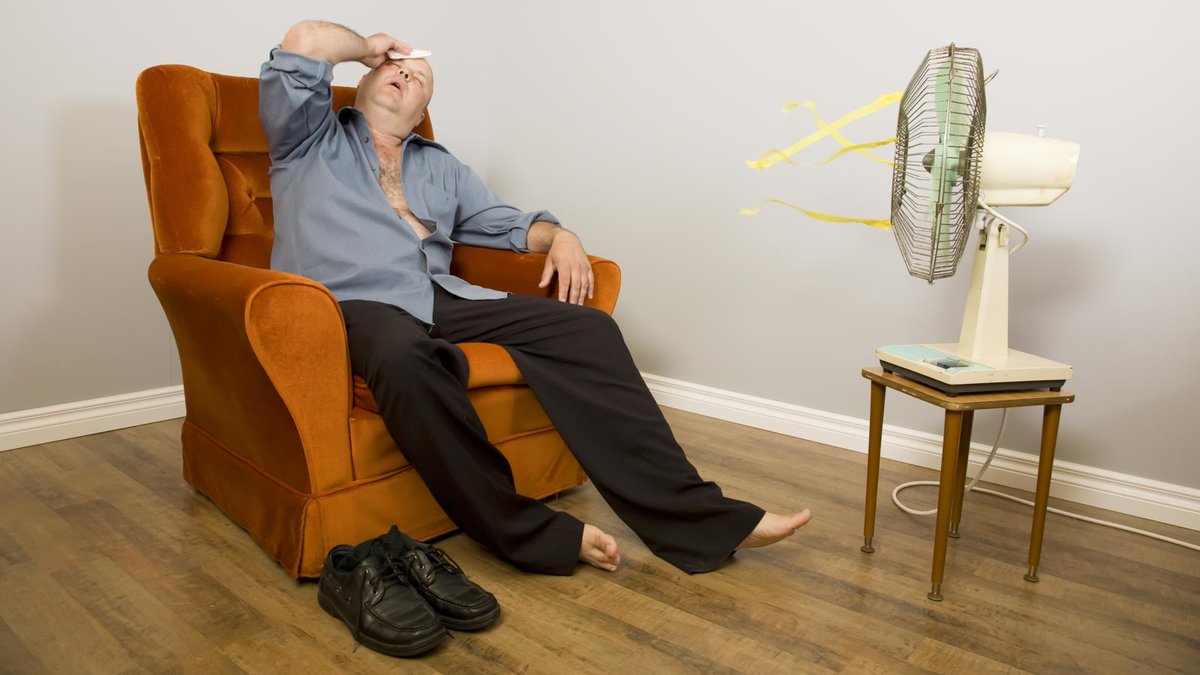 Mann auf einem Sessel sitzt vor einem Ventilator und leidet unter der Hitze im Raum