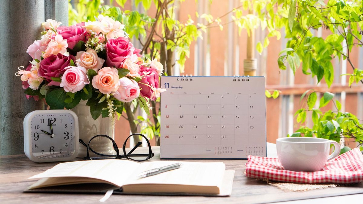 Kalender auf einem Tisch mit Notizbuch, Tasse, Wecker und Blumenstrauß