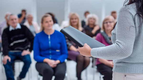 Referentin mit Ordner in Ihrer Hand spricht zu (meist älteren) Zuhörern