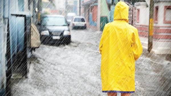 Person mit gelbem Regenmantel steht in Starkregen und überschwemmter Straße it dem Rücken zum Betrachter iim