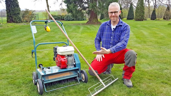 Gartenberater Philippe Dahlmann neben einem blauen Vertikutierer und einer Rasenharke, die auf dem Rasen stehen.