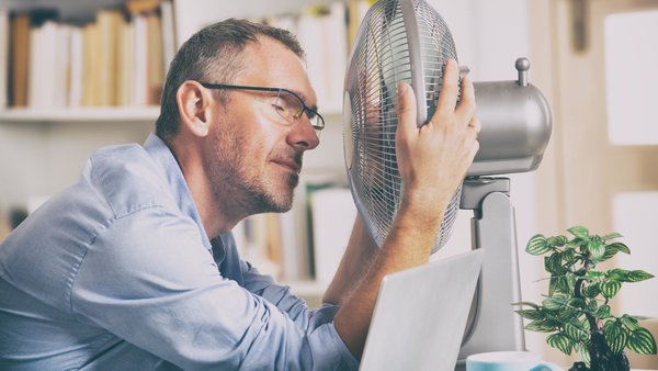 Schwitzender Mann sitzt zuhause ganz nah mit dem Gesicht vor einem Ventilator