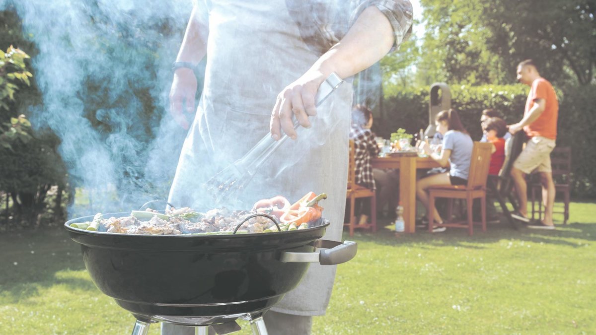 Mann am Grill im Vordergrund, im Hintergrund Gruppe im Garten