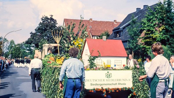 Mitglieder der Gemeinschaft Rheda laufen bei einem Festumzug mit einem blumengeschmückten Wagen durch den Ort.