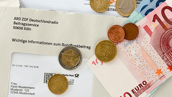 Briefumschlag mit Adresse für Rundfunkbeitrag, bedeckt von Euro-Münzen und Scheinen