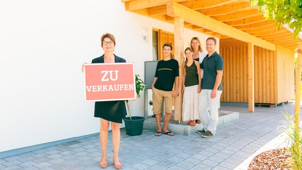 Maklerin mit ZU verkaufen-Schild vor einer jungen Familie am Hauseingang