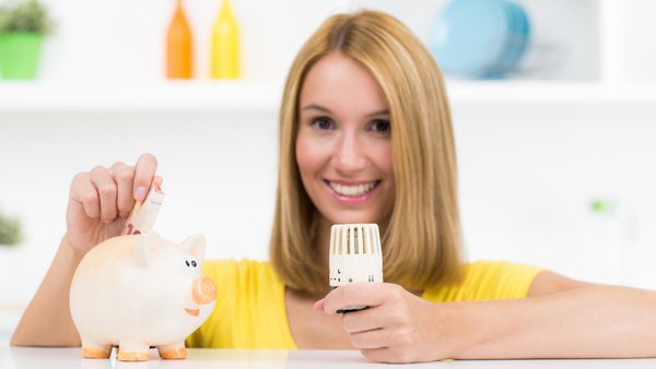 Lächelnde junge Frau mit Heizungsthermostat in der Hand steckt Geldschein in ein Sparschwein