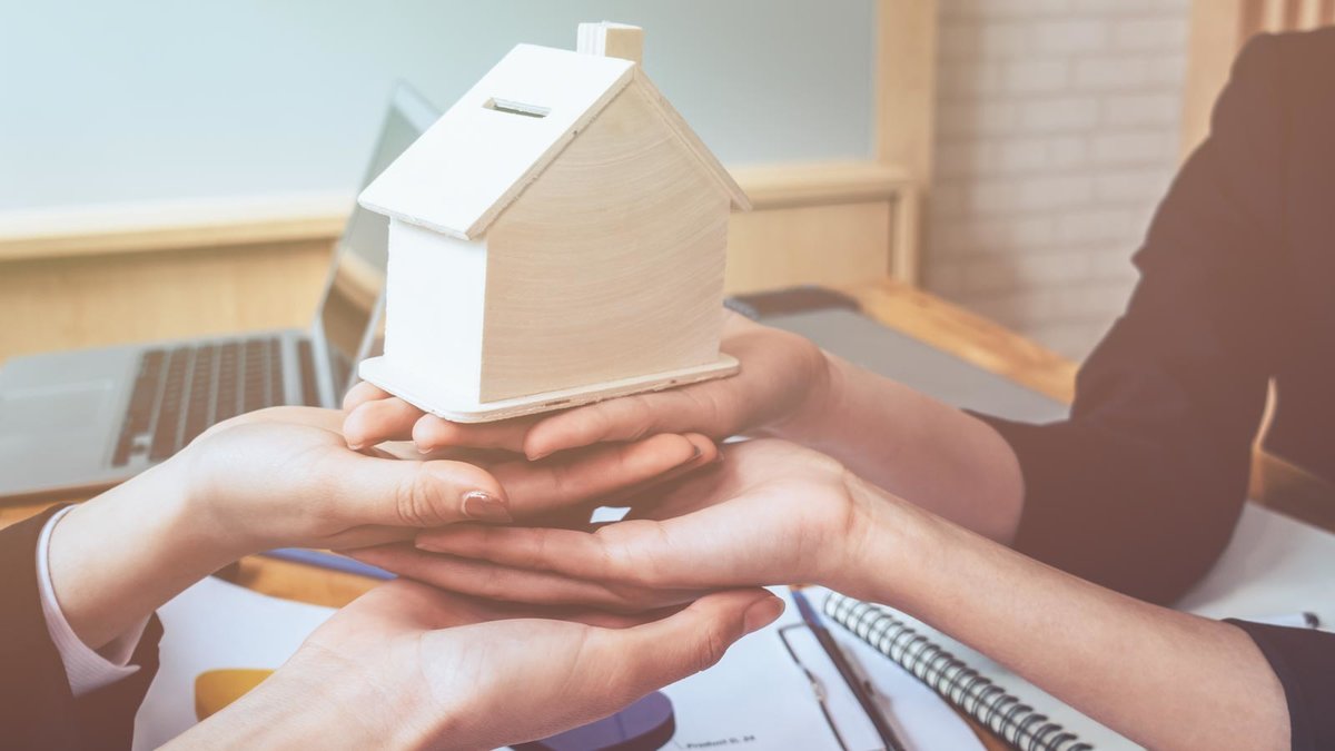 Übereinandergelegte Hände tragen ein Modellhaus aus Holz