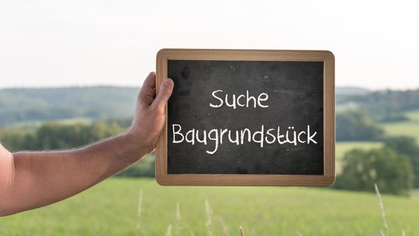 Schild mit der Aufschrift "Suche Baugrundstück" wird vor dem Hintergrund einer Landschaft ins Bild gehalten