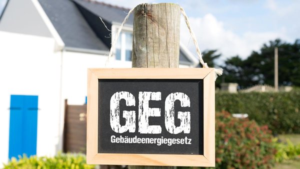 Holzpfahl mit einen Schild und der Aufschrift GEG Gebäudeenergiegesetz vor einem Einfamilienhaus im Hintergrund