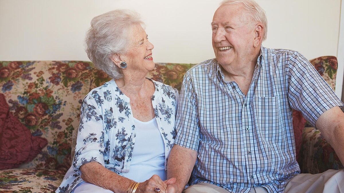 Älteres Paar sitzt händchenhaltend auf dem Sofa und lächelt sich an